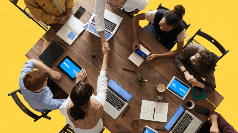 Vue plongeante d’un groupe de professionnels collaborant autour d’une grande table en espace de co-working, avec ordinateurs, tablettes et carnets de notes.
