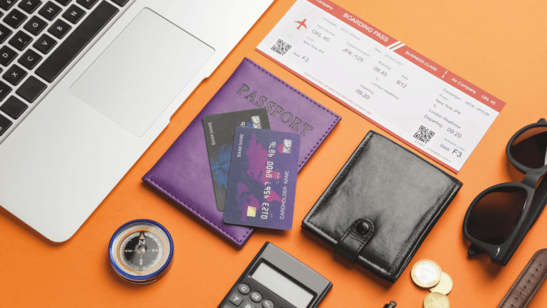 Vue de dessus d’objets essentiels pour un voyage d’affaires : ordinateur, passeport, cartes bancaires, billet d’avion, portefeuille, boussole, lunettes et pièces de monnaie sur fond orange.