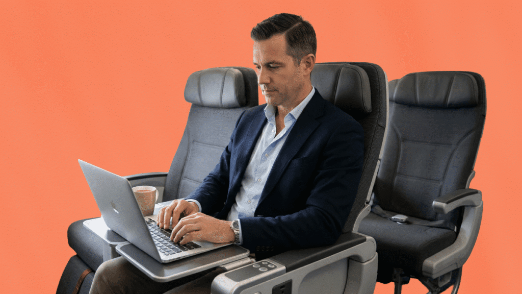 Photographie d'un homme en costume travaillant sur un ordinateur portable, confortablement installé dans un siège spacieux, illustrant l'application d'une politique voyages classe avion optimisée. Il est assis dans une cabine premium sur un fond orange uni, une tasse posée sur sa tablette, symbolisant la productivité durant les déplacements professionnels.