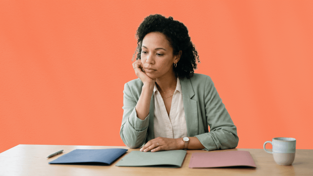 Une professionnelle assise à son bureau, la tête dans la main, hésitant entre trois dossiers de couleurs différentes pour choisir son agence de voyages d'affaires sur un fond orange.